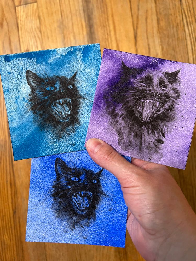 Photo of a hand holding three small square pieces of art: black and white yowling cats on textured watercolor backgrounds in purple and blue.