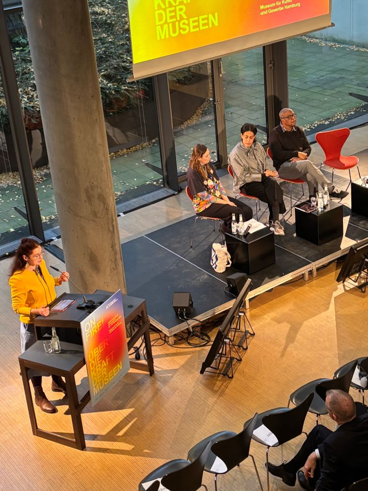 Man sieht eine Frau am Redepult und das Podium, bestehend aus drei Personen. 
