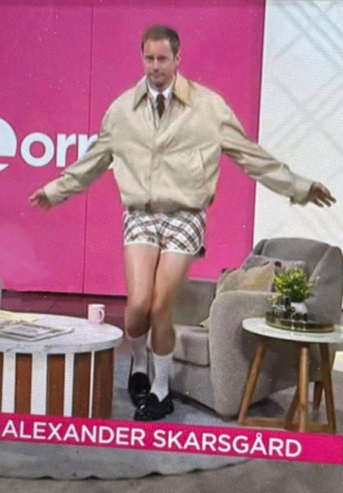 Alexander Skarsgård on Lorraine. He's standing up, arms floppily outstretched, wearing a short beige jacket zipped up, brown tie, white collared shirt, brown and white plaid short shorts and black loafers with white ankle socks.