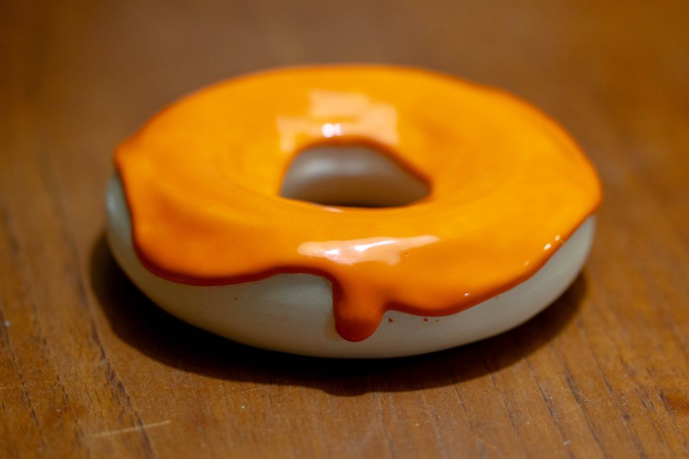 2024-05-09 Toronto Ontario: Colour photo of a ceramic donut. A bright orange glazed icing lays atop a bright white bare clay 'donut'  sitting on a red wooden surface.