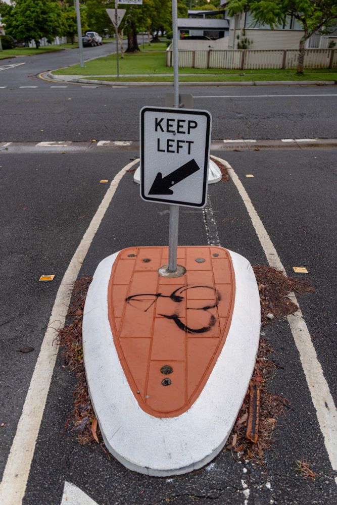 2025-01-18 Brisbane, Australia - A colour photo looking down at a traffic median containing a road sign 'KEEP LEFT'. The red brick facade of the ground surface has a crude black spray painted phallic also pointing to the left lane similar to the road sign.

Canon R6 28mm f2.8 1/25s iso400