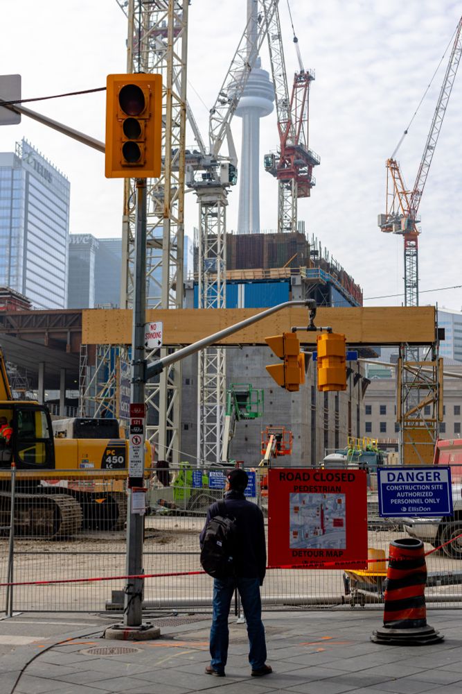 2022-10-30 Toronto, Ontario: Colour photo of a man standing at the edge of younge street blocked by a metal fence with a sign reading 'Road Closed, Pedestrian Detour Map' with a large chaotic construction site with multiple cranes and a concrete structure slowly blocking the view of the CN tower.

Canon R6 50mm f5 1/400sec iso100