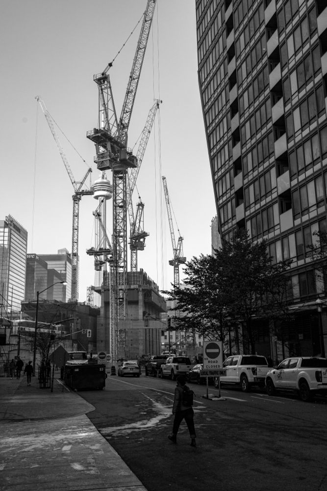 2022-11-04 Toronto Ontario Canada - Black and White portrait street photo looking down the esplanade street towards the CN Tower. The view being blocked by 5 construction cranes and a growing concrete pillar of a new building.  Several 'road closed' signs line the street towards the construction with a single person crossing in mid-stride.

Canon R6 35mm f7.1 1/100sec iso160