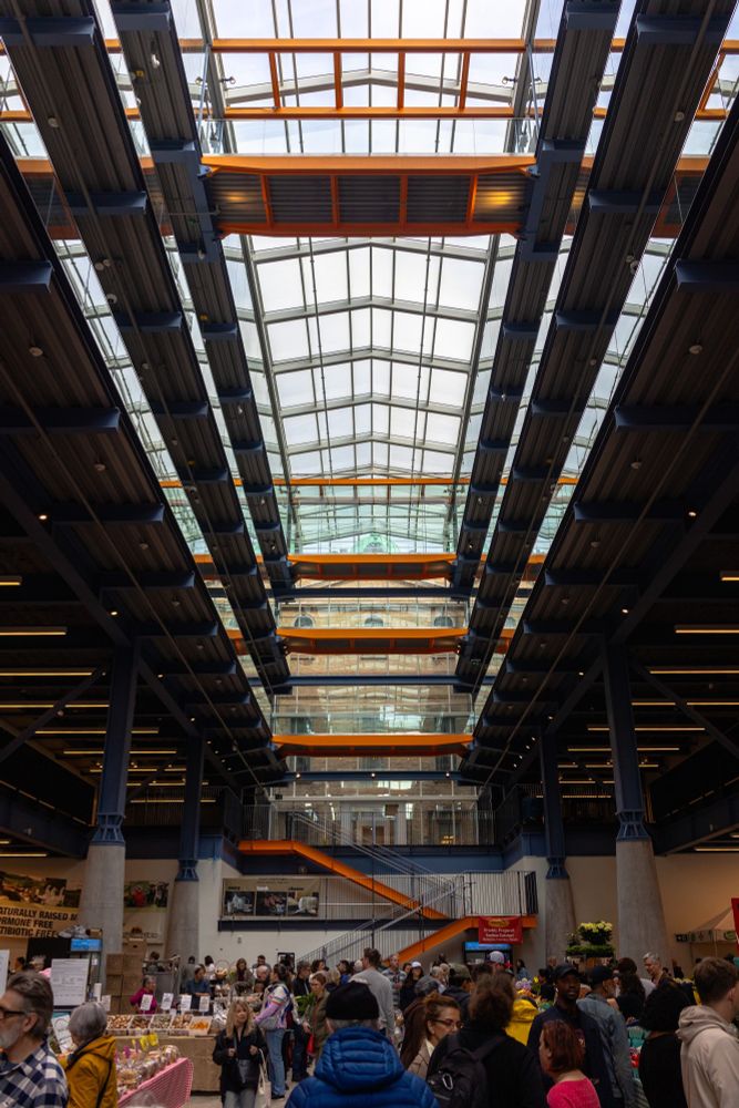 2025-04-19 Toronto, Ontario, Canada - portrait color photo looking up in the newly built North St Lawrence Market building with large metal pillar lining the sides of a massive skylight in the center of the building. Crowds fill the hall, while bright orange highlights the traversable stairs and bridges in the center of the building.

Canon R6 28mm f3.5 1/640s iso400