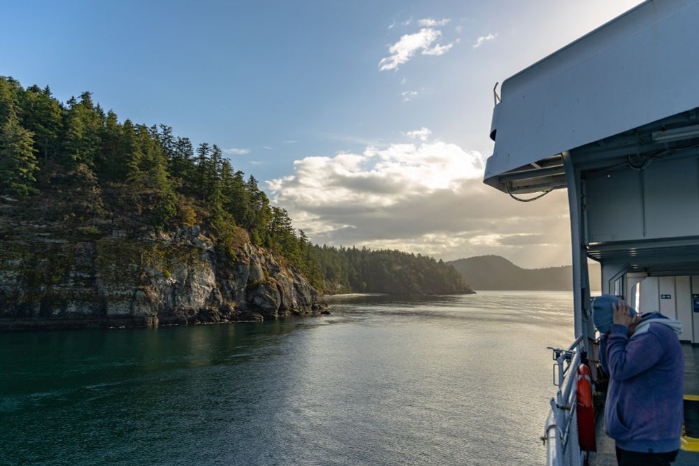 2021-09-18 Vancouver Island British Columbia Canada - Color photo taken in transit to Vancouver Island looking out over the water of a narrow passage. Large rock cliff line the shores with big green coniferous tree forest covering the top. On the ship, a man in purple hoodie and blue cap stand cupping his ear with hands tightly as though to block an immense sound. 

Canon R 24mm f4 1/1250sec iso100