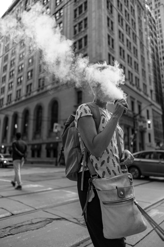 2022-09-12 Toronto, Ontario: Black and white photo of short haired lady crossing King st in the mid afternoon. She is exhaling a large cottony cloud from a vape obscures her face.

Canon R6 24mm f1.8 1/800sec iso125