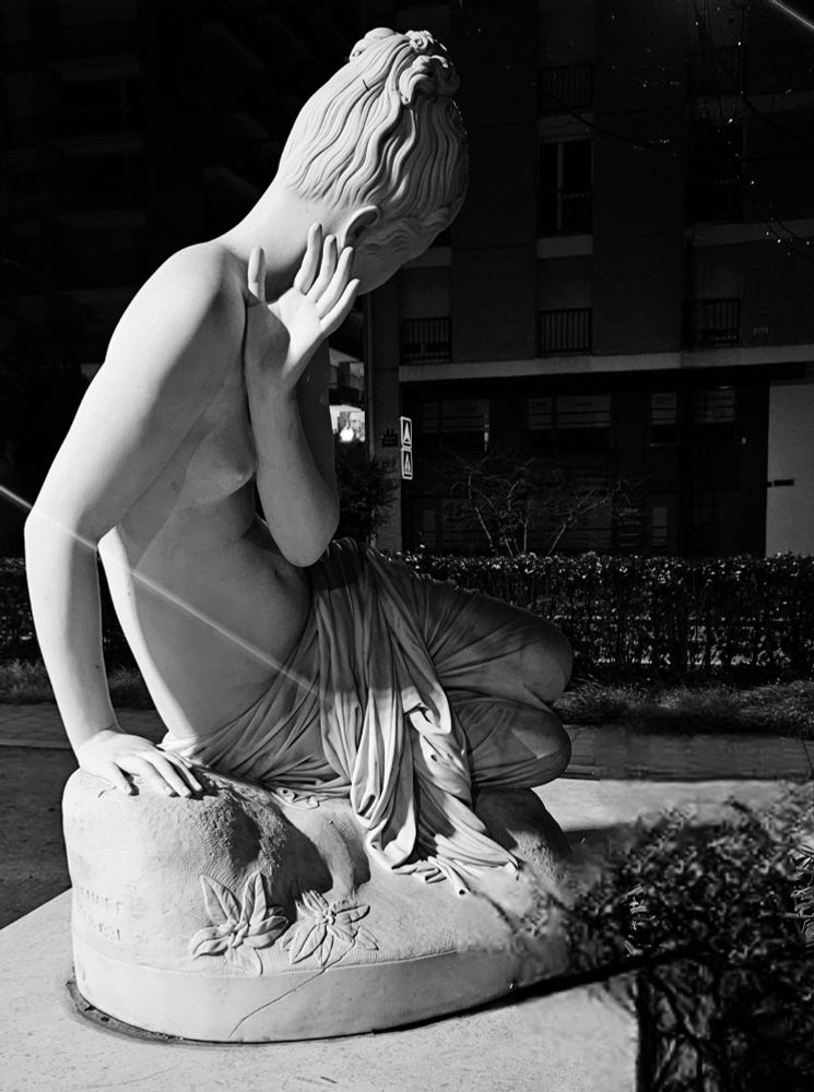 Statut blanche, en ville, d'une femme de profil, torse nue, qui se cache le visage de la lumière venant de gauche