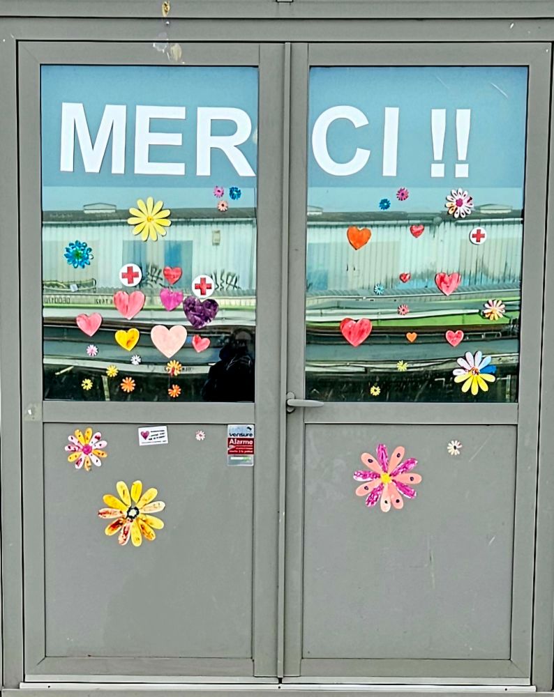 Porte d'extérieur d'un bâtiment, sur le canal, en verre et metal, où il est écrit "Merci" en autocollant blanc, en dessous, des cœurs et fleurs autocollants sur le verre de la porte. 