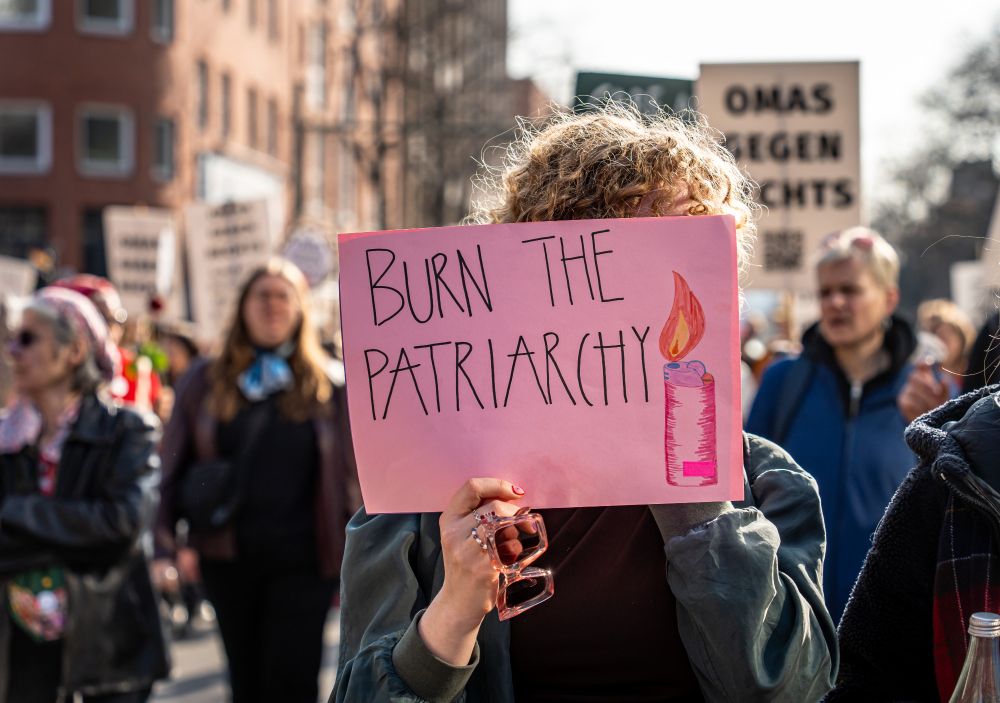 Eine Frau hält sich ein rosa Papier vors Gesicht auf dem ein Feuerzeug gemalt ist und "Burn the Patriarchy".
