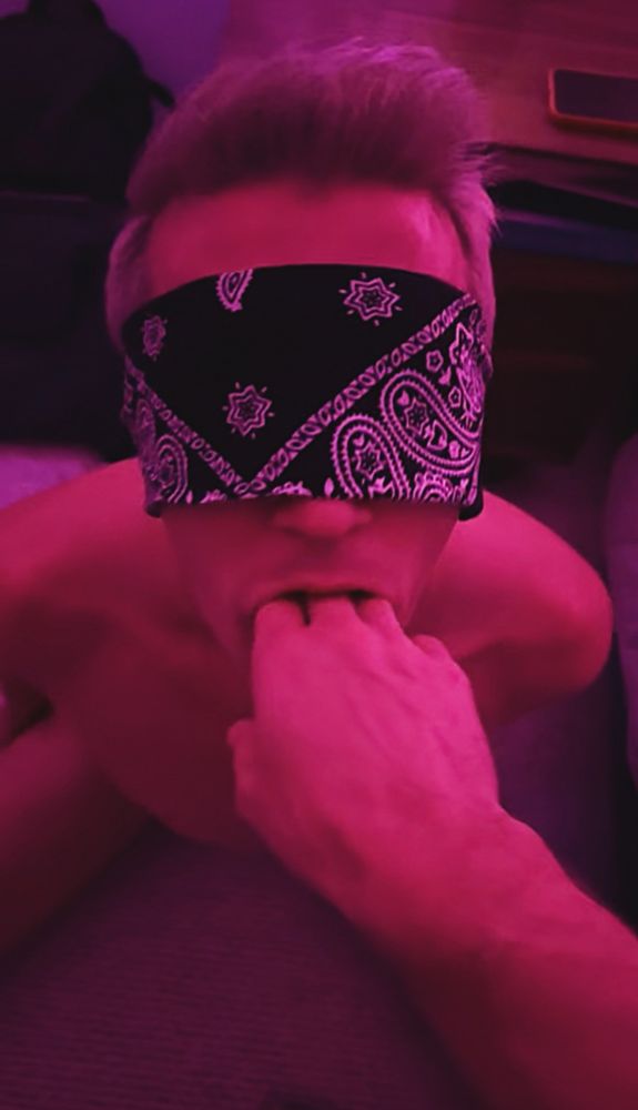 Boy on his knees, wearing only a jockstrap and blindfold, under pink lighting. Someone’s hand is stuffing 3 fingers in his mouth. 
