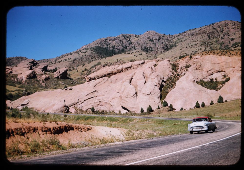 A vintage Kodachrome slide showing a road scene from the American West in the 1960s; the road goes to the right and then up foothills to the left, and a car is shown in the foreground heading towards the viewer