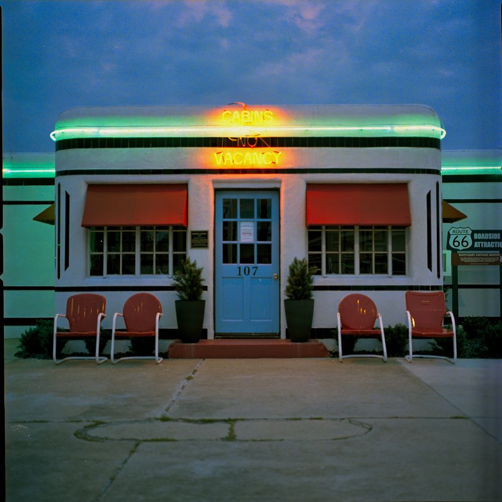 A square photograph shows the front of an art deco style motel office at dusk, the black and white building has red awnings and other accents, and it is wrapped with vintage neon
