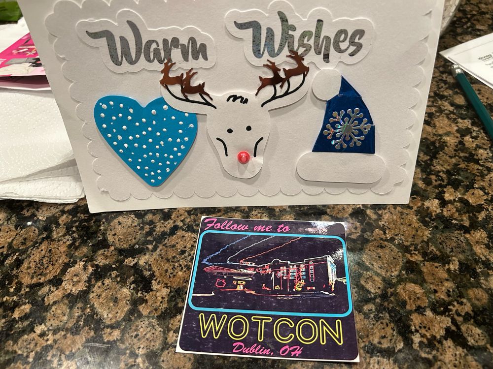 A cute homemade card with a blue heart with white polka dots, a  white reindeer with a red nose and a blue/white Santa hat with a sparkling snowflake. Below it is an exclusive sticker with the WOTCon hotel with a psychedelic filter with text that reads, “Follow me to WOTCon, Dublin, OH.” Credit: LewsJunior