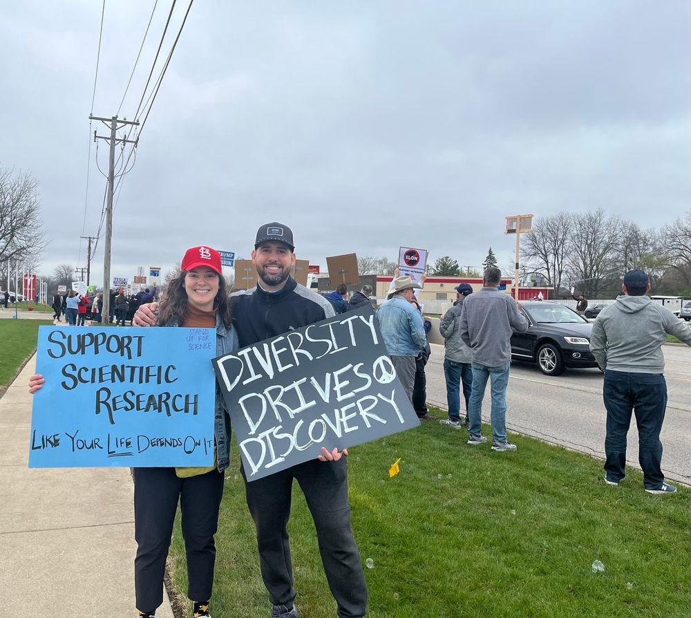 Laura in a red STL hat smiling holding a blue sign that says “Support scientific research like your life depends on it” and she is next to her brother who is wearing a black hat, smiling and holding a black sign that says “diversity drives discovery” in white paint. There is a peace sign on the poster. Behind them is a long sidewalk with many people with signs all standing along a main road