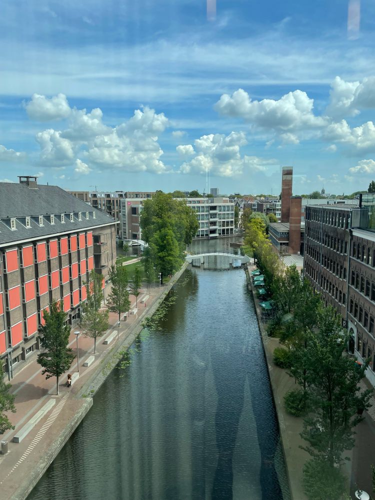 View from ‘de Brug’ (the bridge) on University of Amsterdam campus, there is a canal below, with 5 story red brick buildings on either side, in the distance is a white bridge and in the far distance you can see a windmill. It’s a sunny day with some clouds. 