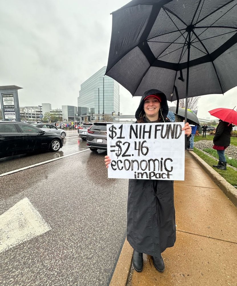 Laura standing with a white sign with black text: “$1 NIH FUND=$2.46 economic impact” she is on a sidewalk next to a busy road, holding an umbrella, in the background are more people with signs. She is wearing a red STL hat, a full body length rain jacket, and black boots. She is smiling because she is proud to be standing up for science 