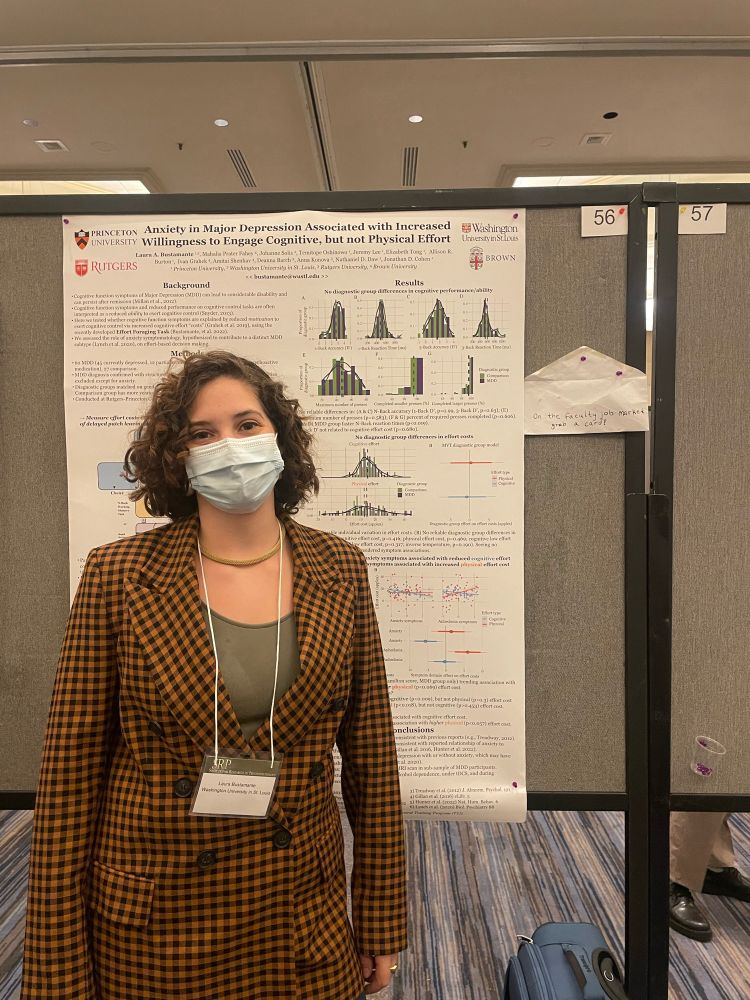 Laura presenting a poster at the society for research on psychopathology. She has a poster board behind her, she is wearing an orange blazer with black checkering, an olive green shirt, a gold necklace, a blue mask, and a name tag.
