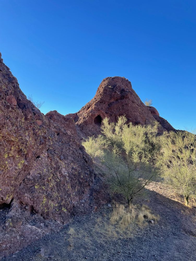 Image taken at camelback mountain, the sky is blue, the foreground has some desert bushes, and there are two rocks jutting out of the ground. The rock on the right has a hole in it, maybe something is living there!