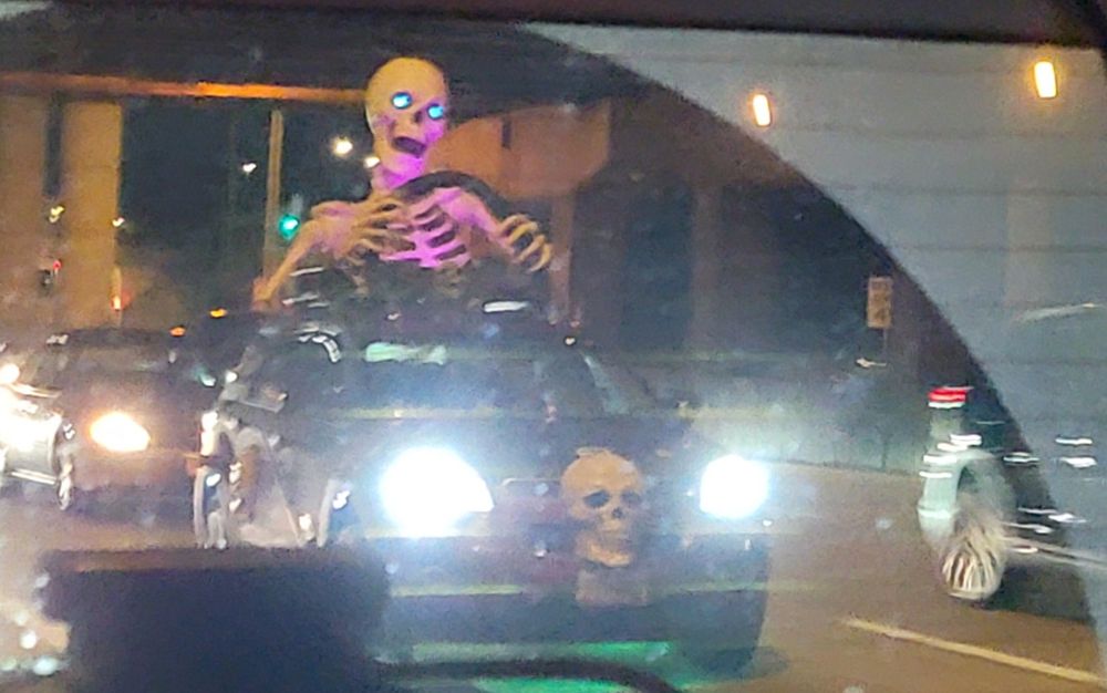 Skeleton with blue glowing eyes driving a car. There is a skull in the grill of the car as well. 