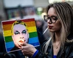 A woman with cigarette in her mouth also lights poster of Victor Orban (in drag) on pride flag background