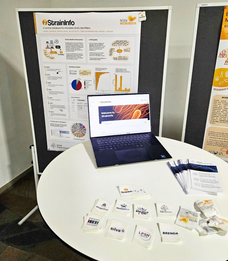 Photo of the StrainInfo poster at the NFDI4Microbiota conference in Leipzig. In front of it stands a table with a laptop opened to the StrainInfo website and promotional material 