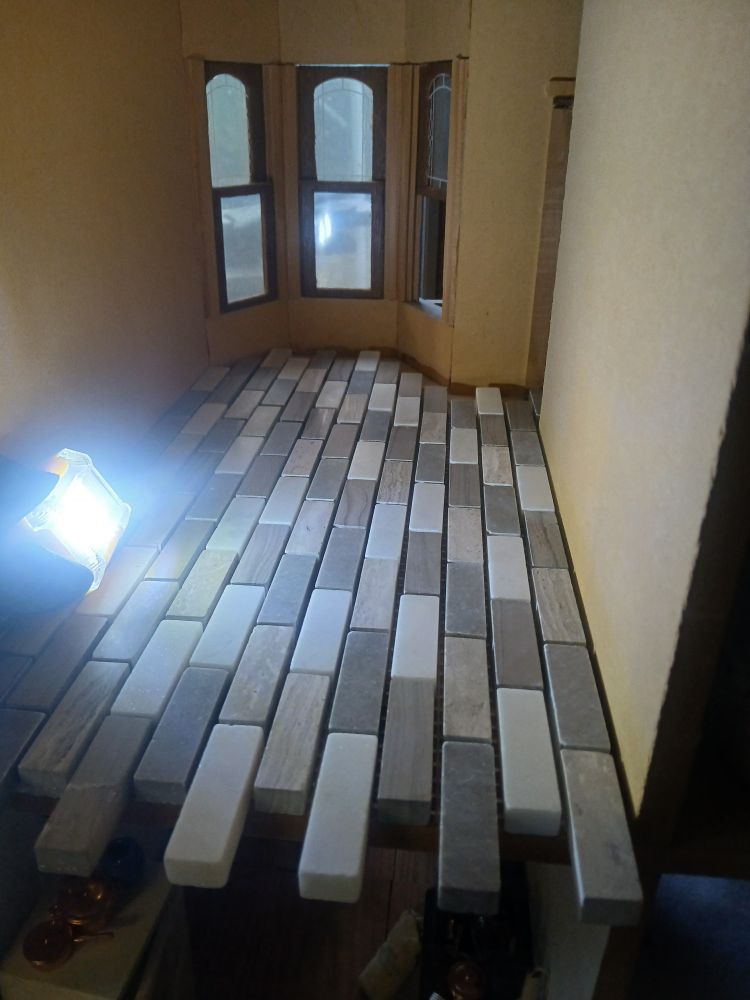 An empty room in a dollhouse. The walls are painted white and a set of mixed white and grey tiles covers the floor. There are windows on the far side and a headlamp lights the space.