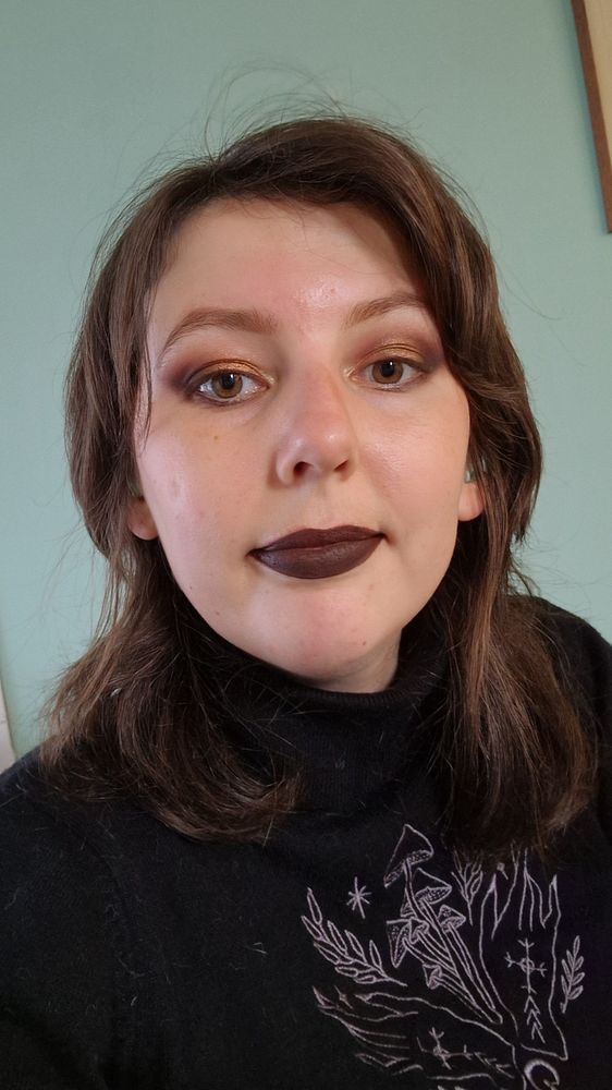 Photo of me, a white nb person with shoulder-length brown hair. I'm wearing dark brown lipstick with matching dark eyeshadow. 