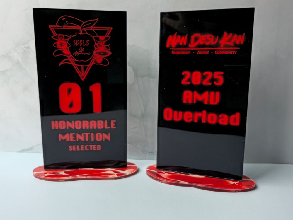 Two acrylic awards.
They look like the nerv seele monoliths from evangelion. 
The one on the left has a parody image of the seele logo that says "seele of approval" 01 honorable mention selected
The one on the right has the logo for nan desu kan
2025 amv overload