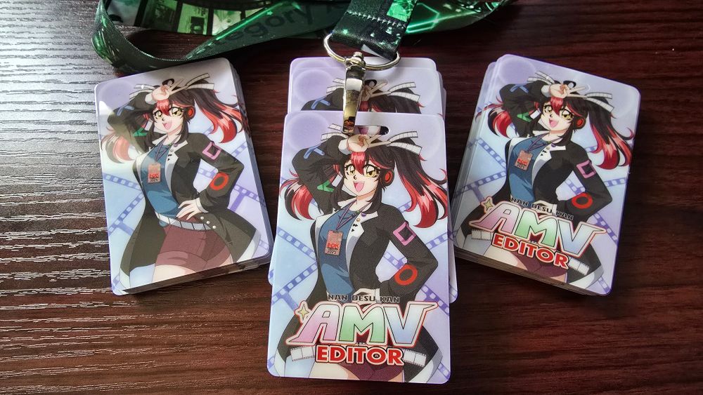 3 stacks of AMV badges. They have a mascot character doing a sailor moon inspired pose, and the text "Nan Desu Kan AMV Editor"
The middle badge is on a green lanyard.