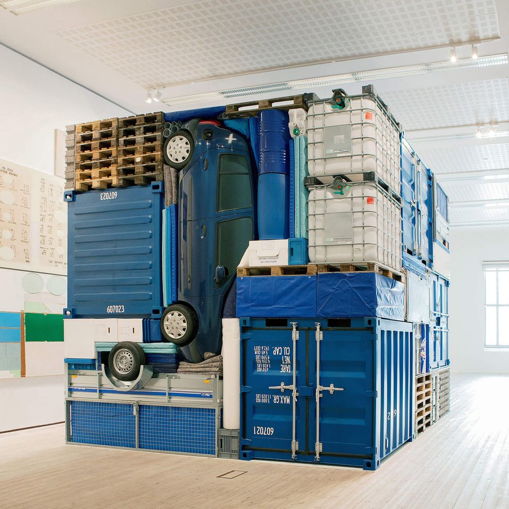 The installation appears as a large, perfectly geometrical cube composed entirely of various blue items, including: 
Shipping containers
A car
Pallets
Barrels
Large plastic liquid containers