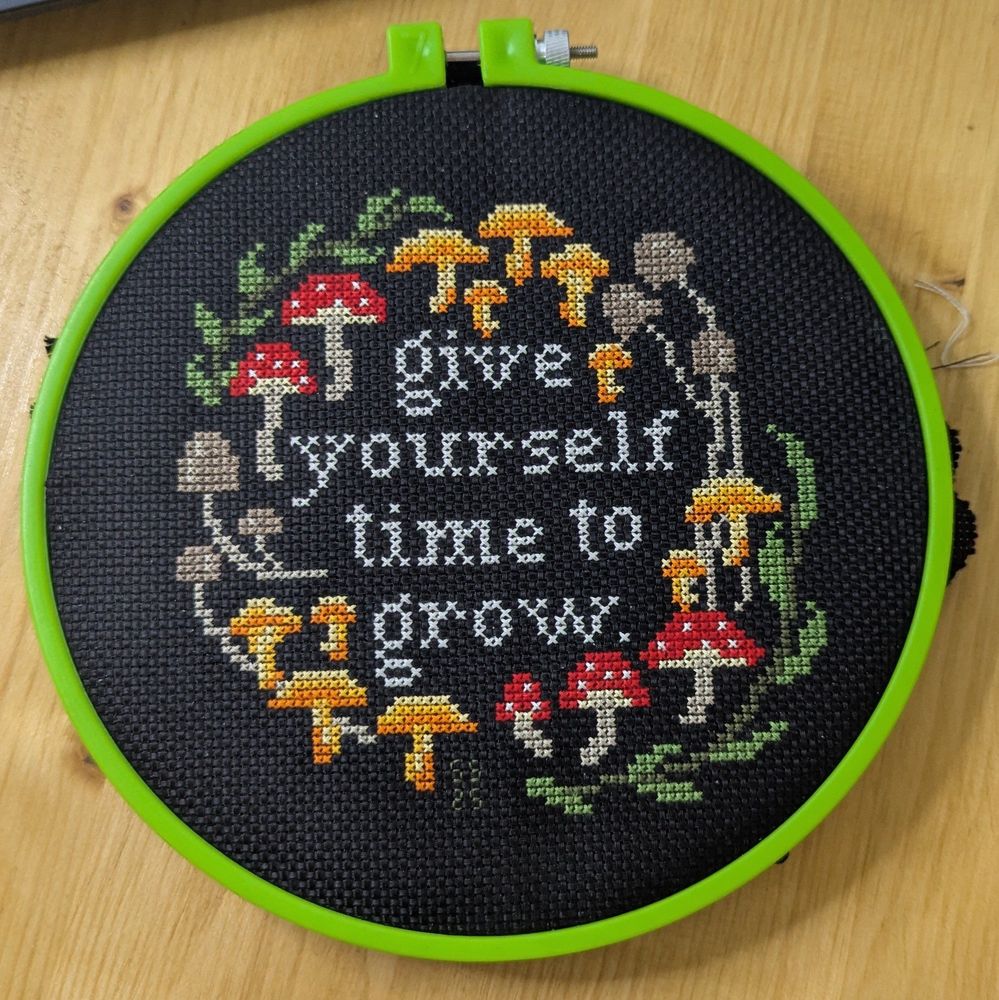 Cross stitches mushrooms and greenery surrounding the words "give yourself time to grow" on black Aida cloth. 