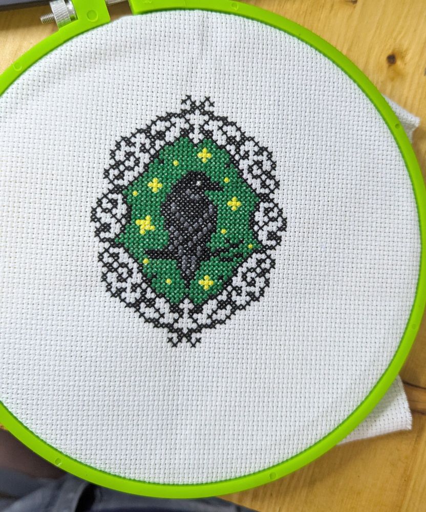 Cross stitched raven on a branch on a dark green background. There are yellow stars as well. The border looks like black scroll work, all on white Aida fabric. 
