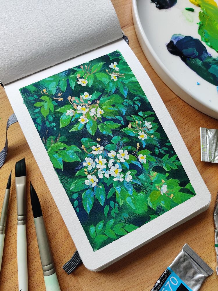 Photo shows a sketchbook laying open on a desk. The book is open to a page, displaying a painting of green leaves and white flowers. Paintbrushes, gouache paint tubes and a palette surround the painting. 
