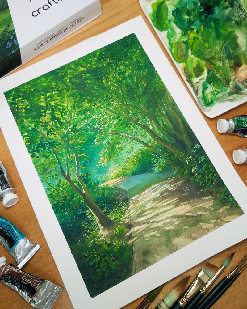 Gouache painting sitting on a wooden desk, surrounded by paint supplies such as brushes, a palette, and paint tubes. The painting is of a pathway through trees on a sunny summer day, with foliage on either side of the path. The pathway is in partial shade, with dappled light illuminating some areas. 