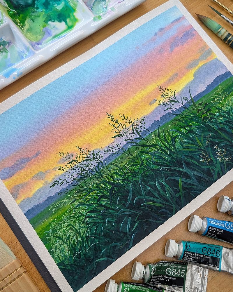 The photo shows a gouache painting on a desk, surrounded by paint supplies. The painting is of a summer sunset over a field with tall grasses. 