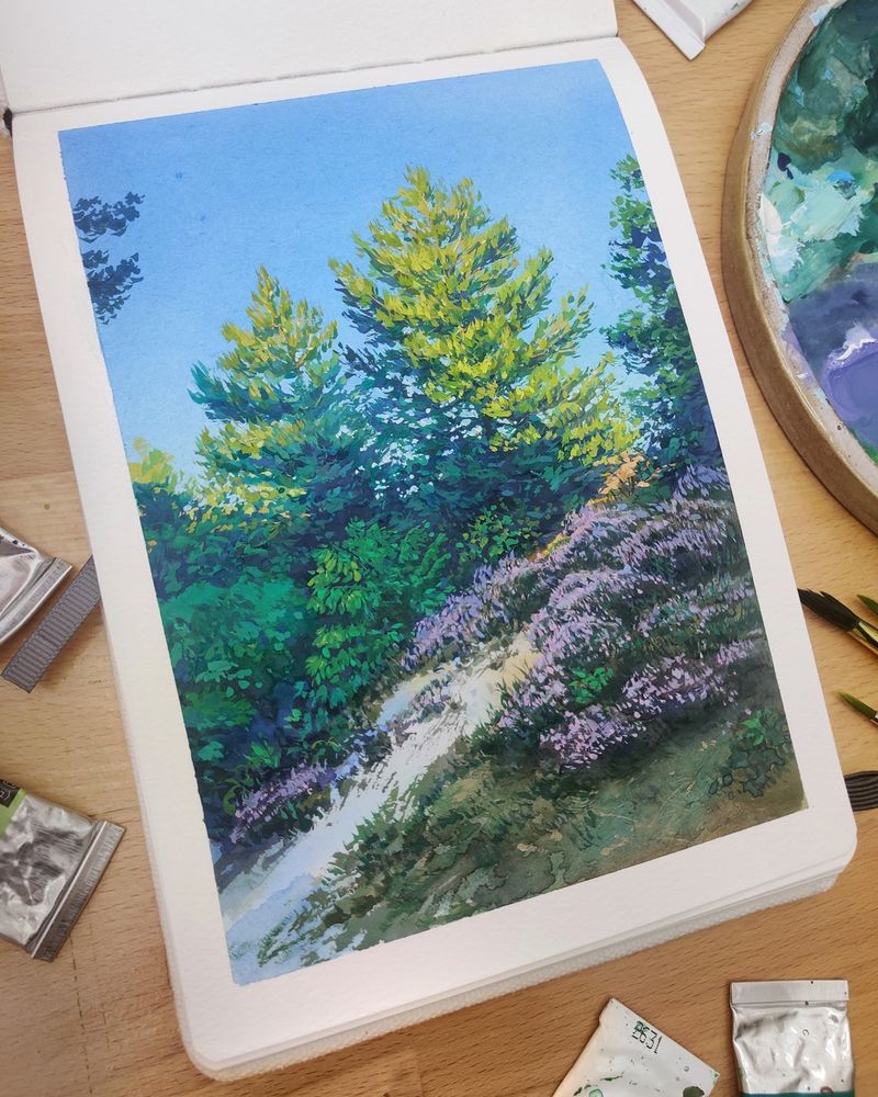 Photo shows a sketchbook open on a wooden desk. The sketchbook is open to a page, displaying a gouache painting of pine trees and heather atop a sandy hill. Paint tubes, brushes and a palette surround the book. 
