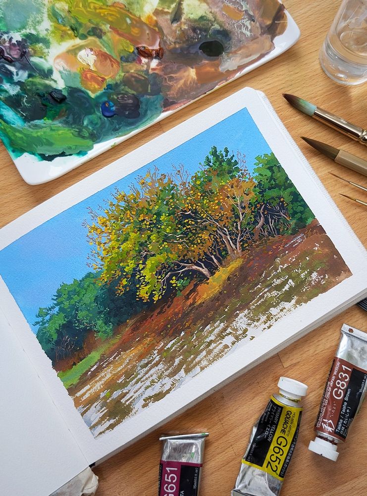 A photo of a watercolor sketchbook open on a desk, surrounded by a palette, paintbrushes and gouache paint tubes. The painting is of a small tree surrounded by other foliage in the Autumn.