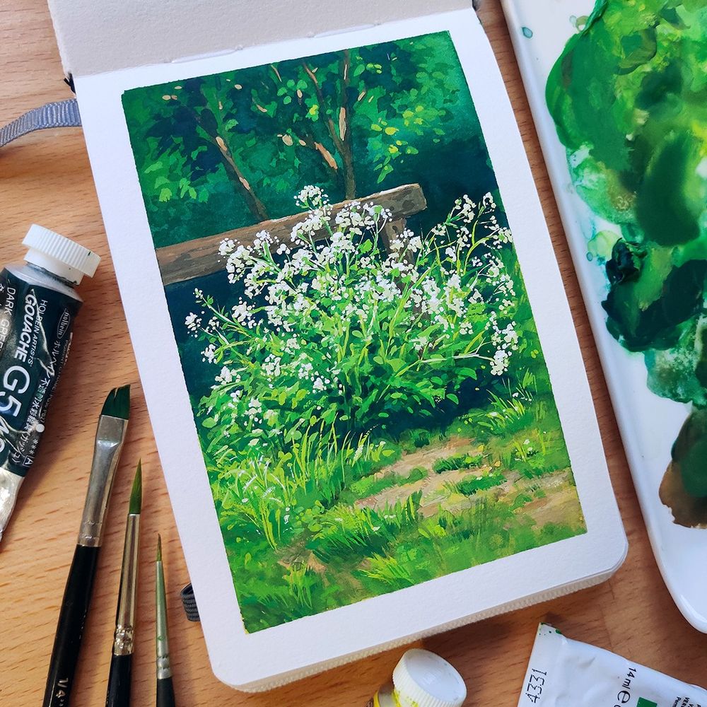 Photo of a small sketchbook laying open on a wooden desk, surrounded by paintbrushes, gouache paint tubes and a paint palette. The open page shows a painting of a nature scene. There is a white flowering plant in front of a fencepost with trees and foliage behind. 