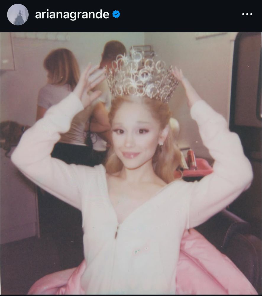 a photo from ariana grande’s recent post that features her in a light pink sweater and bubble crown, her hands are steadying it on her head