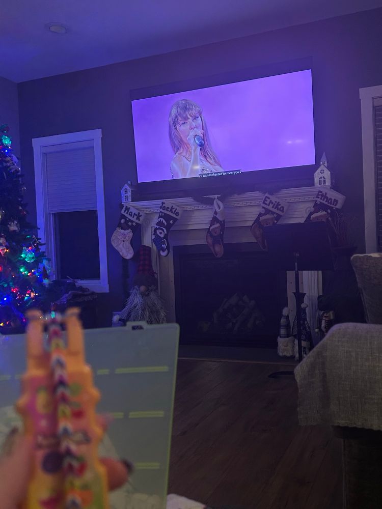 a picture of taylor swift during the speak now era on the eras tour with the lyrics “i was enchanted to meet you” on the screen. also seen are some stockings, a christmas tree, and my looming tool which i’m making an eras rainbow loom bracelet on