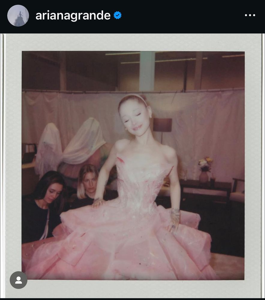 a photo from ariana grande’s recent post in her pink “no one mourns the wicked” bubble dress, at what looks like a dress fitting. two other women are in the bottom left hand corner of the photo examining the dress