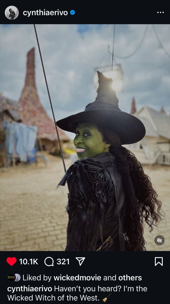 a behind the scenes photo posted by cynthia erivo of her in her wicked part 2/movie 2 wicked witch costume and green skin with the caption “haven’t you heard? i’m the wicked witch of the west. 🧹” presumably on the munchkinland set, as yellow bricks and the munchkin hut sets can be seen in the background.