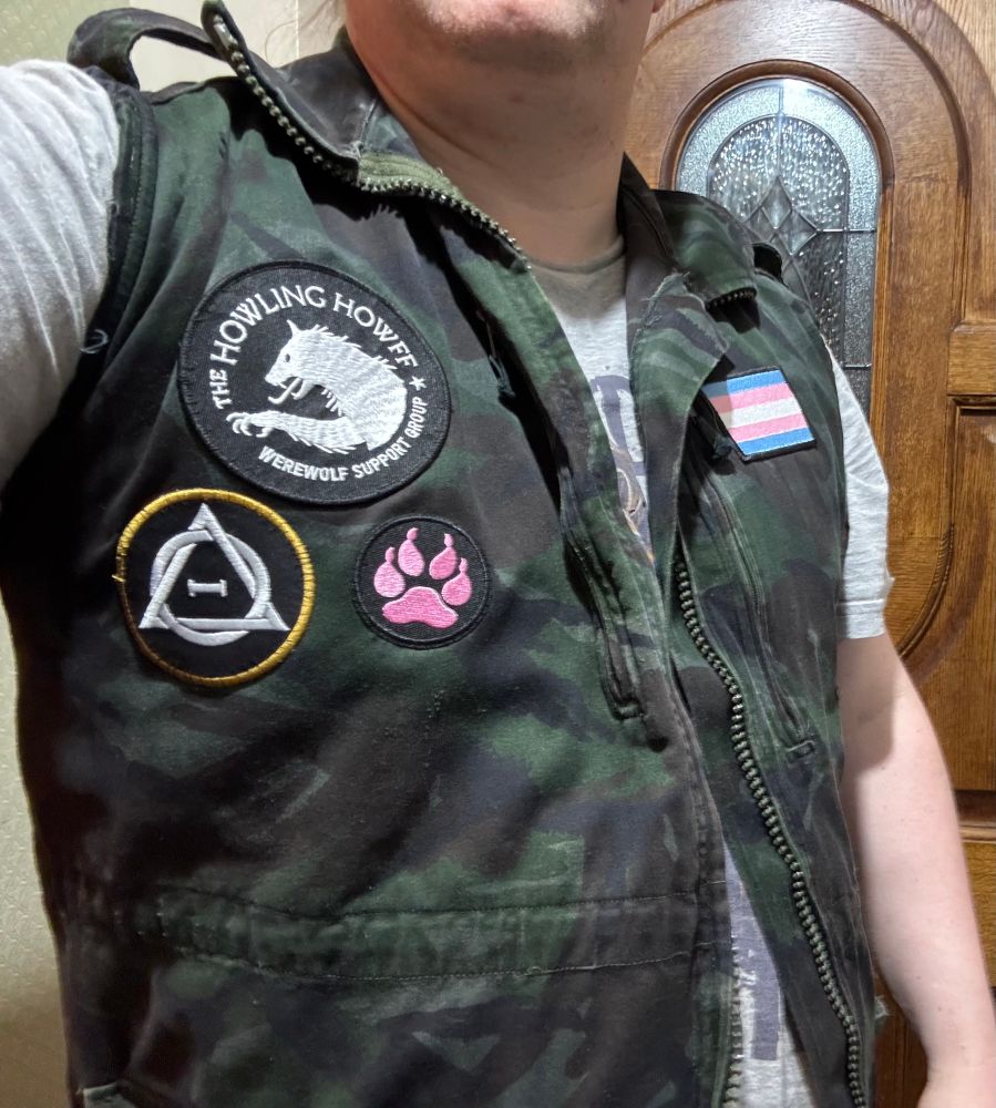 A sleeveless camo jacket dyed black, which makes the colours subdued, and has some patches on it. On one side is a patch of a werewolf with the words "The Howling Howff Werewolf Support Group", a Therian Theta Delta patch, and a pink paw patch. On the other side is a trans flag. 