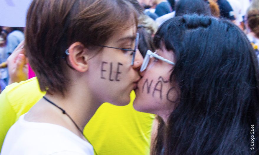 Duas mulheres se beijam. Na face de uma está escrita a palavra "ele" e na da outra a palavra "não" 