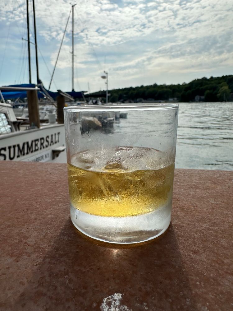 A glass of light yellow liquor with ice.  In the background is a row of boats- one motor boat and several sailboats.  The sky is slightly overcast, and the water is light gray.