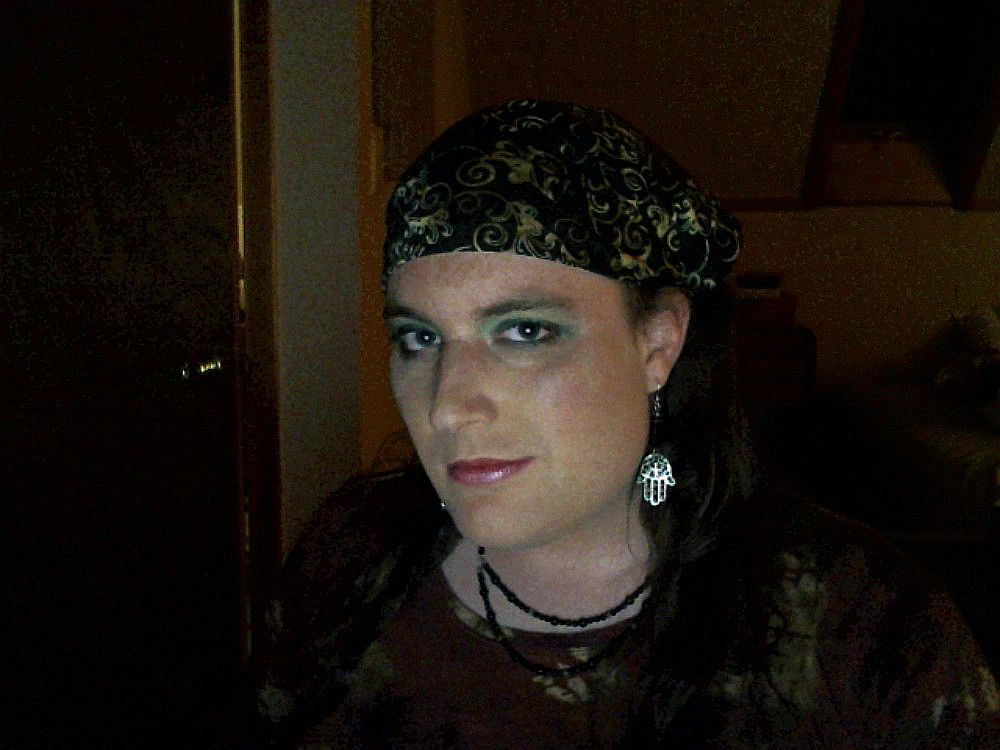 A young light skinned woman with long dark hair tied back in a black and gold batik dyed bandana, wearing green eye makeup, glossy plum lipstick, silver hamsa earrings, a layered necklace made of black beads, and an earth toned top. She has piercing eyes, looking directly at the viewer, with a sly grin, facing slightly away from the camera so as to better display both her lovely dangling earrings and her saucy attitude. She is illuminated by a soft, warm, single light source, in what appears to be an attic bedroom with a slightly ajar door on the left.