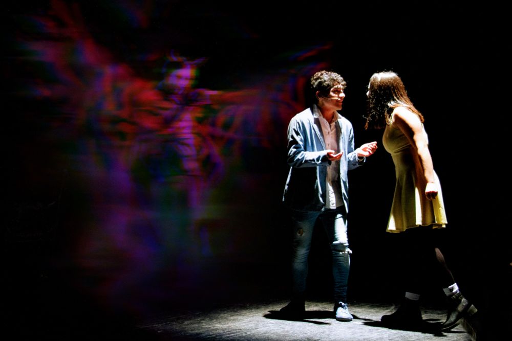Two actors performing under a stage light. Behind them, a blurry figure spreads what appear to be wings. It is formed by red, blue and green blurs. One of the actor's feet is teetering off the edge of the stage.