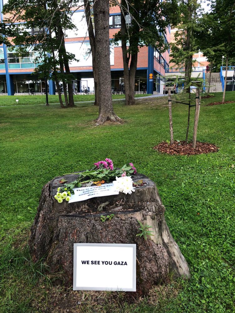 An arrangement with flowers and signs in front of the University of Jyväskylä. One saying "We see you Gaza," the other one saying "This is a space for tribute and solidarity with Gaza."