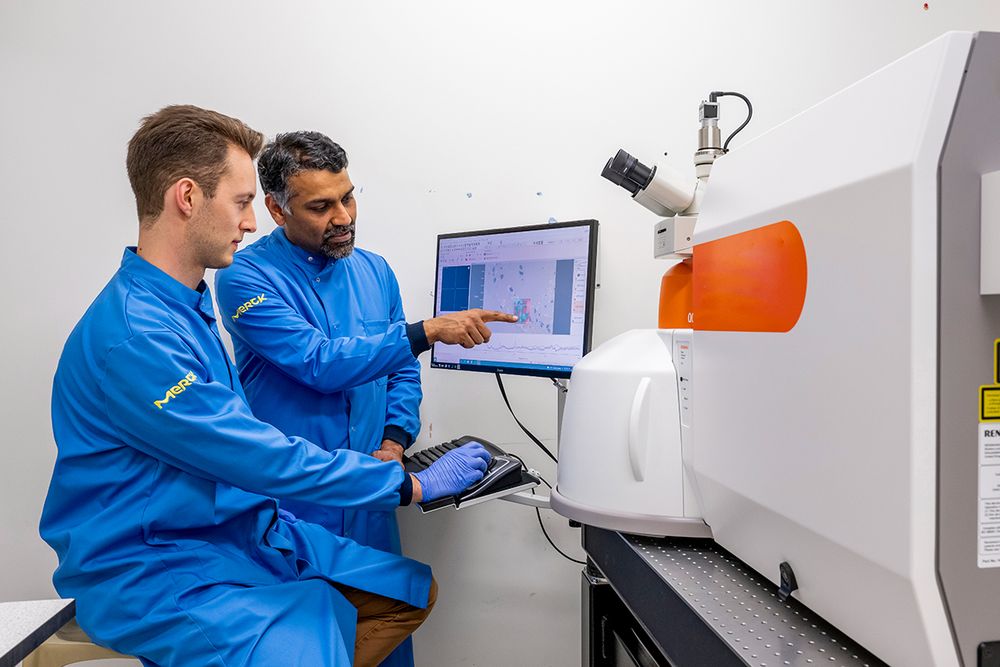 Two scientists in blue lab coats are working in a laboratory. One is seated, using a keyboard, and the other is standing and pointing at data on a computer screen. They are both focused on the monitor.