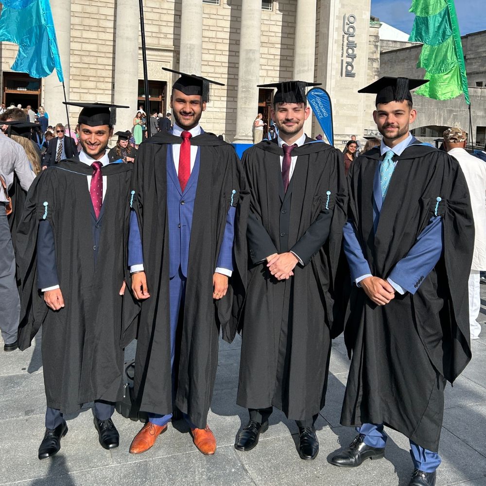 Four individuals in graduation gowns and caps stand in front of the Guildhall, under a blue sky. Colourful flags are visible on either side.